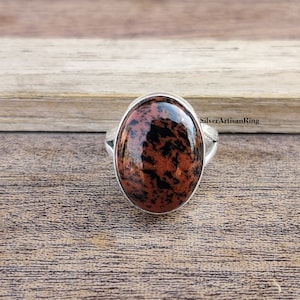Puede incluir: Anillo ovalado con una piedra pulida de color marrón rojizo con inclusiones negras, engastada en una banda plateada. El anillo tiene una superficie lisa y brillante y se muestra sobre una superficie de madera. El texto "SilverArtisanRing" es visible.