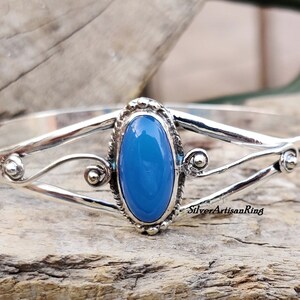 K&ouml;nnte beinhalten: Ein silbernes Armkettchen mit einem blauen ovalen Edelstein, der in der Mitte eingesetzt ist. Das Armband hat ein zartes, filigranes Design mit Schn&ouml;rkeln und kleinen Perlen.