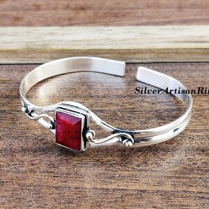 May include: A silver bangle bracelet featuring a red gemstone set in a square bezel. The bracelet has a delicate, wavy design and is open on one side.