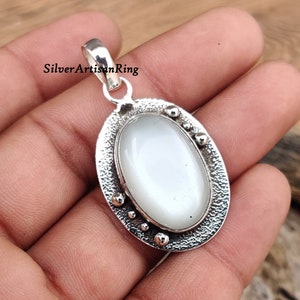 Op de afbeelding: Een zilveren hanger met een witte ovale edelsteen gezet in een getextureerd ovaal frame. De hanger heeft een lus om aan een ketting te bevestigen.
