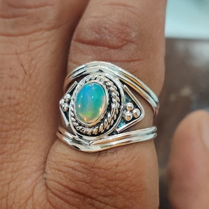 May include: A silver ring with a large opal gemstone set in a decorative band. The ring has a detailed design with small beads and a rope-like border around the stone.