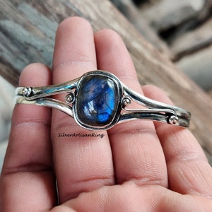 Puede incluir: Un brazalete de plata con una piedra preciosa de labradorita azul. El brazalete tiene un dise&ntilde;o sencillo con dos bandas y un peque&ntilde;o acento de cuentas a cada lado de la piedra preciosa.