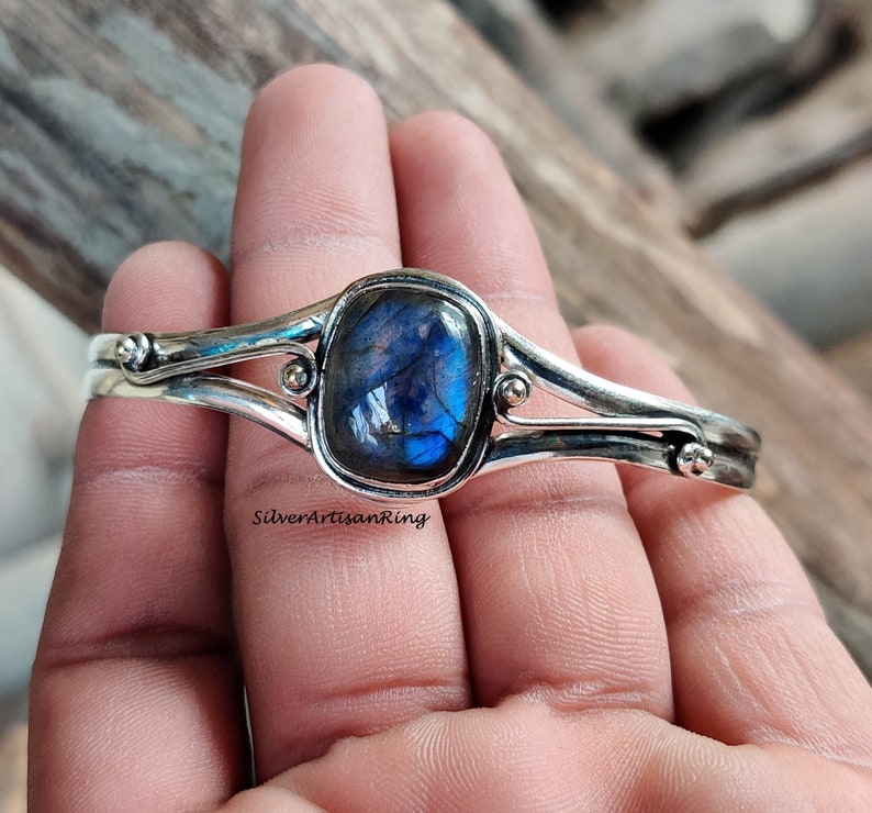 Puede incluir: Un brazalete de plata con una piedra preciosa de labradorita azul. El brazalete tiene un dise&ntilde;o sencillo con dos bandas y un peque&ntilde;o acento de cuentas a cada lado de la piedra preciosa.