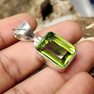 Pode incluir: Um pingente retangular com uma pedra preciosa verde brilhante emoldurada em uma estrutura de metal prateado. A pedra preciosa tem uma superf&iacute;cie facetada e o pingente tem uma argola para uma corrente. O pingente &eacute; segurado em uma m&atilde;o.