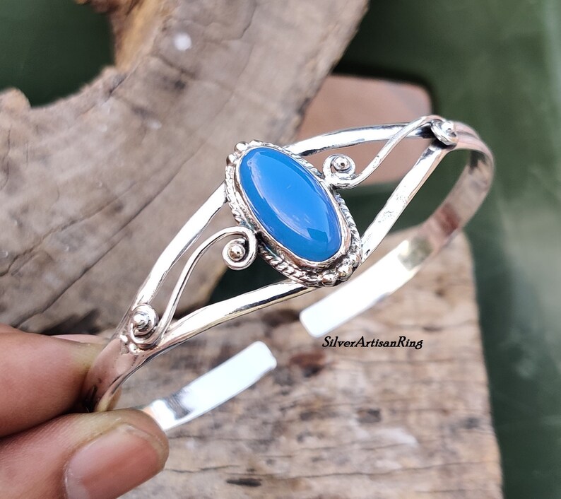 K&ouml;nnte beinhalten: Ein silbernes Armreif-Armband mit einem hellblauen ovalen Edelstein, der in einer silbernen Fassung sitzt. Das Armband hat ein zartes, verziertes Design mit Spiralen und kleinen Perlen.