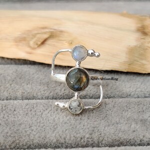 Könnte beinhalten: Ein silberner Ring mit drei Steinen. Der mittlere Stein ist ein großer, schillernder Labradorit. Die beiden kleineren Steine sind Mondsteine. Der Ring ist im modernen Stil mit einem gebogenen Band gestaltet.