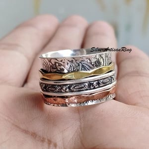 May include: A silver spinning ring with four bands. The top band is silver with a textured design, the second band is gold, the third band is silver with a textured design, and the bottom band is copper.