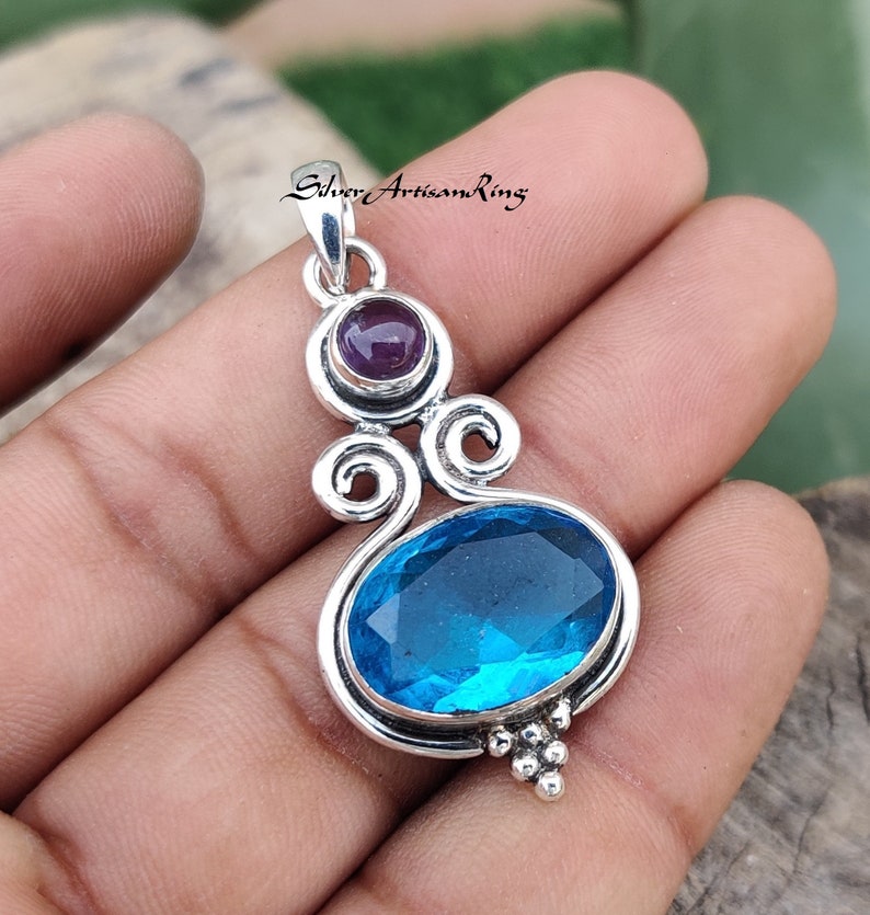 May include: A silver pendant with a blue topaz gemstone and a smaller amethyst gemstone. The pendant has a delicate, ornate design with swirls and a beaded bottom.