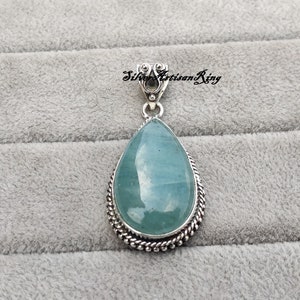 May include: A teardrop-shaped pendant with a light blue gemstone set in a silver frame. The pendant has a silver bail for attaching to a chain.