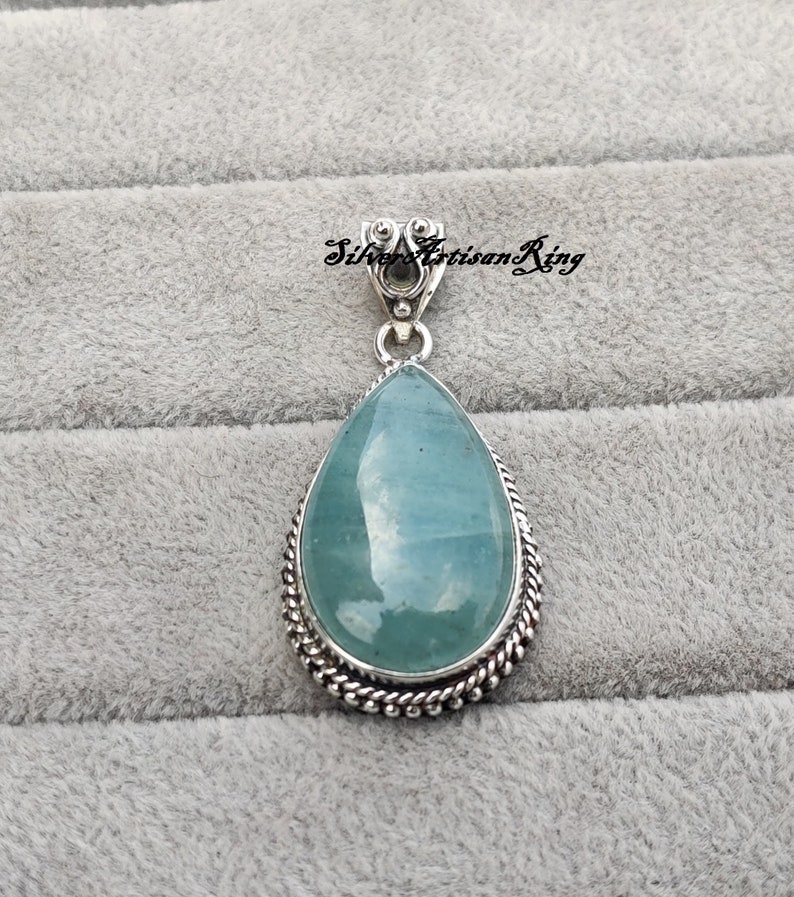 May include: A teardrop-shaped pendant with a light blue gemstone set in a silver frame. The pendant has a silver bail for attaching to a chain.