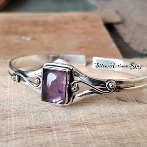 Puede incluir: Una pulsera de plata con una piedra preciosa púrpura engastada en un marco cuadrado. La pulsera tiene un diseño delicado y curvo con pequeñas bolas decorativas.