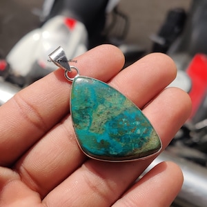 Könnte beinhalten: Ein tropfenförmiger Anhänger mit einem grünen und blauen Edelstein, der in Sterlingsilber gefasst ist. Der Edelstein hat ein geflecktes Muster aus Grün, Blau und Braun.
