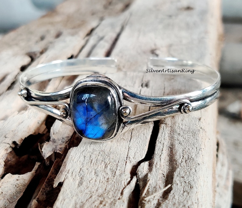 Puede incluir: Un brazalete de plata con una piedra preciosa de labradorita azul engastada en un engaste de bisel. El brazalete tiene un dise&ntilde;o delicado con dos bandas curvas y peque&ntilde;os detalles dorados.