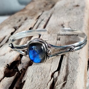 Puede incluir: Un brazalete de plata con una piedra preciosa de labradorita azul engastada en un engaste de plata. El brazalete tiene un dise&ntilde;o delicado y retorcido y presenta peque&ntilde;as cuentas de plata.