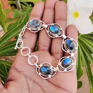 以下が含まれることがあります： 5つの楕円形のラブラドライトの宝石がついたシルバーのブレスレット。各宝石は、装飾的なデザインが施されたシルバーのフレームにセットされています。ブレスレットはトグル留めです。