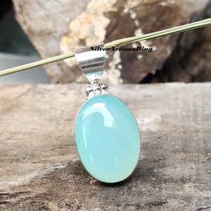 May include: A silver pendant with a large, oval, light blue gemstone. The gemstone is set in a silver bezel and has a smooth, polished surface. The pendant has a simple, elegant design, perfect for everyday wear. The text "SilverArtisanRing" is visible.