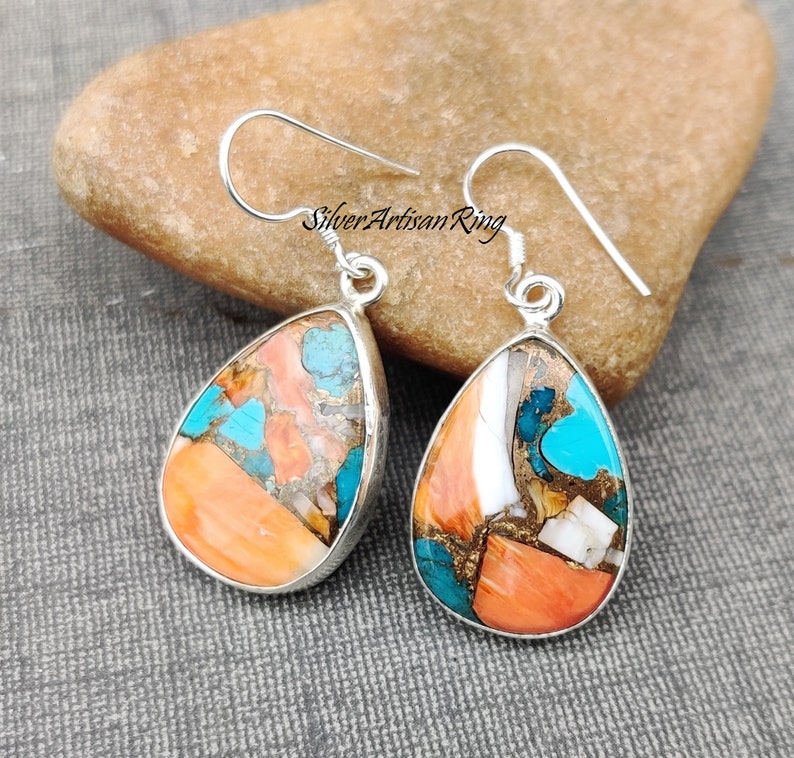 Pu&ograve; includere: Un paio di orecchini a forma di goccia con un gancio d'argento e una pietra multicolore. La pietra &egrave; un mix di turchese, corallo e oro.