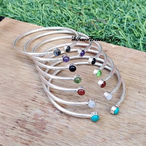 May include: A set of eight silver bangle bracelets with different colored gemstones. Each bracelet has a simple, curved design and a single gemstone at the end. The gemstones are black, amethyst, green, red, white, and turquoise.
