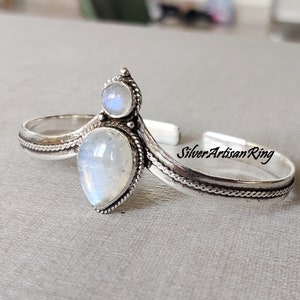 Pode incluir: Uma pulseira de prata com duas pedras de lua. A pulseira tem um design delicado e intrincado com um padrão de corda trançada. O texto "Silver Artisan Ring" está gravado na pulseira.
