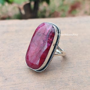 May include: A silver ring featuring a large, rectangular ruby gemstone. The ruby is a deep red hue with internal patterns. The ring has a textured edge and a simple, elegant design. The text "SilverArtisanRing" is visible.