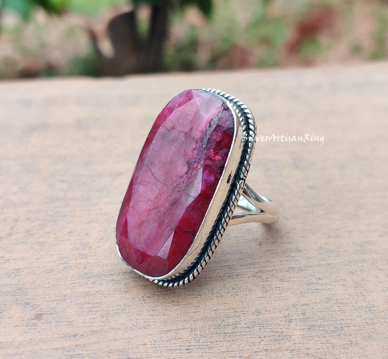 May include: A silver ring featuring a large, rectangular ruby gemstone. The ruby is a deep red hue with internal patterns. The ring has a textured edge and a simple, elegant design. The text "SilverArtisanRing" is visible.