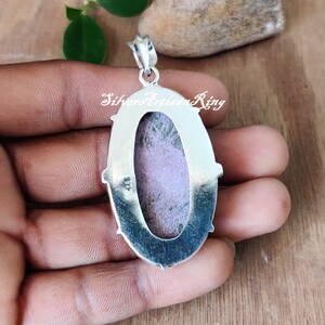 May include: Silver pendant with a purple and green stone set in an oval bezel. The pendant has a silver bail and is on a hand.