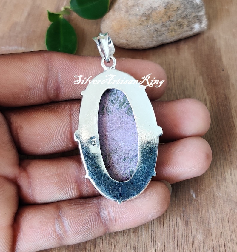 May include: Silver pendant with a purple and green stone set in an oval bezel. The pendant has a silver bail and is on a hand.