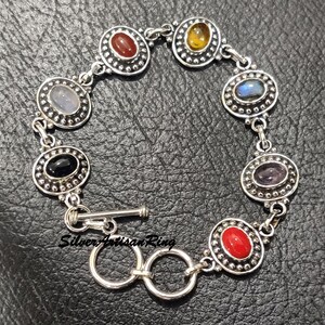 May include: Silver bracelet with seven oval-shaped gemstones set in silver bezels. The gemstones are red, yellow, blue, black, purple, and two shades of blue. The bracelet has a toggle clasp.