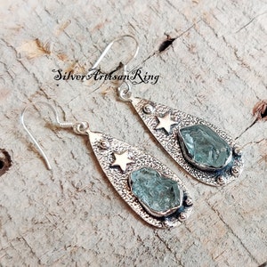 May include: Silver teardrop earrings with a star charm and a light blue aquamarine stone.  The earrings are on a wooden surface.