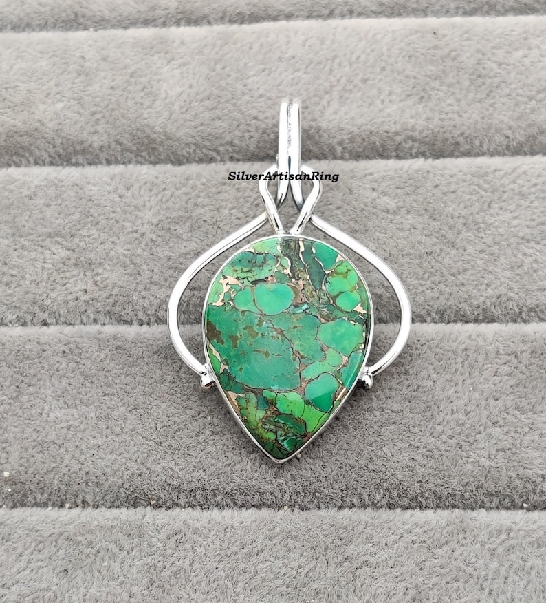 May include: A silver pendant featuring a teardrop-shaped green turquoise stone with brown and white marbling. The stone is set in a silver bail with a loop at the top for attaching a chain.