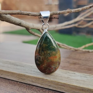 May include: A teardrop-shaped pendant with a green, brown, and orange moss agate stone set in sterling silver. The pendant is hanging from a branch.