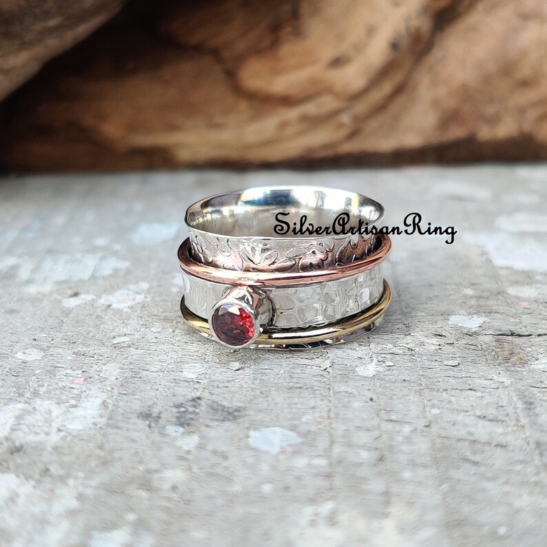 May include: A silver, copper, and gold spinning ring with a red gemstone. The ring is textured and has a hammered finish. "SilverArtisanRing" is written on the ring.