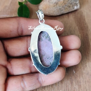 May include: Silver pendant with a purple and green stone set in an oval bezel. The pendant has a silver bail and is on a hand.