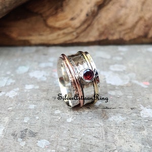 May include: A silver spinning ring with a red gemstone and copper and gold accents. "Silver Artisan Ring" is written on the band.