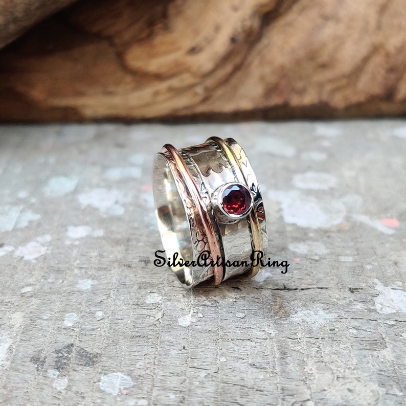 May include: A silver spinning ring with a red gemstone and copper and gold accents. "Silver Artisan Ring" is written on the band.