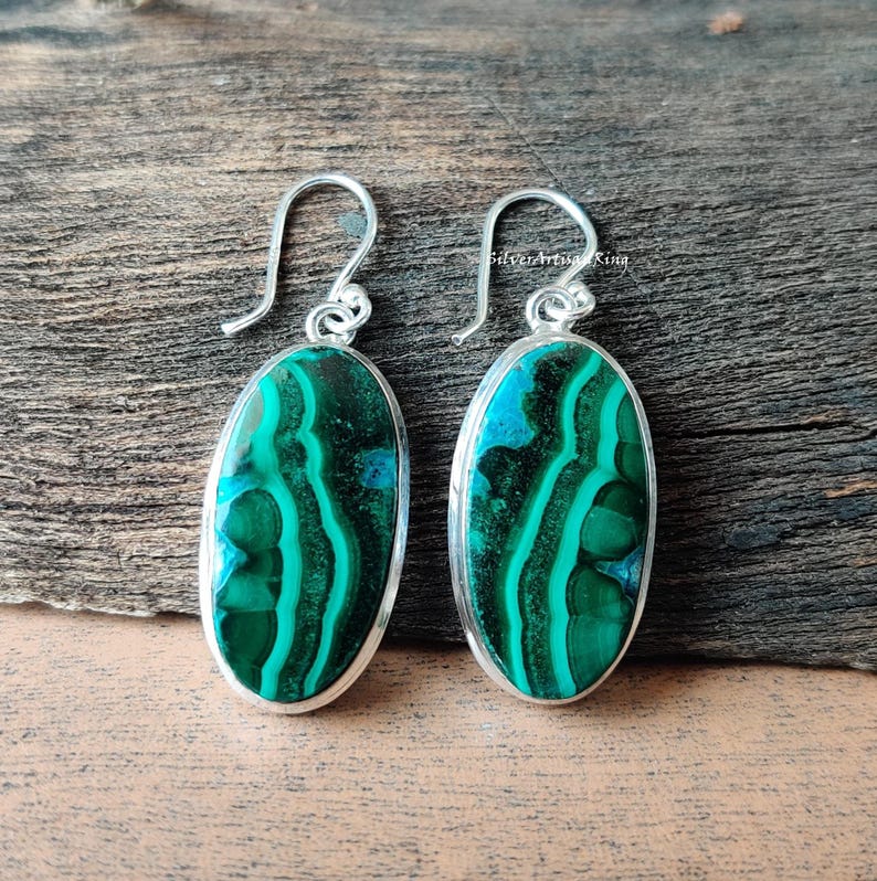 May include: A pair of oval earrings with a silver metal frame. The earrings display a vibrant green and blue malachite stone with distinctive swirling patterns. The earrings are presented on a wooden surface.