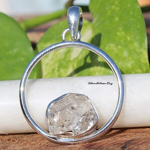 May include: A silver pendant with a clear, faceted crystal stone set in a circle. The pendant is on a white surface with the text "Silverartisan Ring" visible.