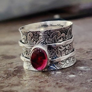 May include: A silver ring with a red gemstone and intricate floral designs. The ring is wide and has a textured surface. "Silver Artisan Ring" is written on the top of the ring.