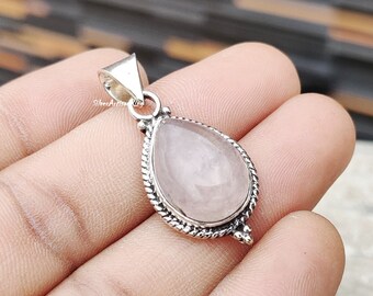 Puede incluir: Un colgante de plata con una piedra de cuarzo rosa en forma de lágrima. El colgante tiene un borde de plata detallado y un aro para sujetar una cadena.