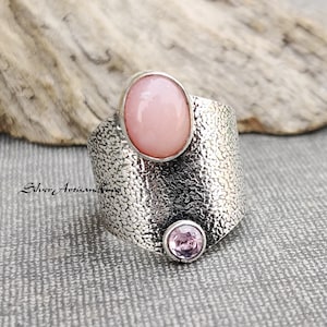 Puede incluir: Un anillo de plata con una banda texturizada y dos piedras preciosas rosas. La piedra más grande es de forma ovalada y la más pequeña es redonda.