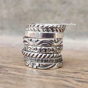 May include: A stack of silver rings with various decorative patterns, including rope, leaf, and scroll designs. The rings are stacked on top of each other, showcasing the different textures and styles. The rings are made of a shiny metal.