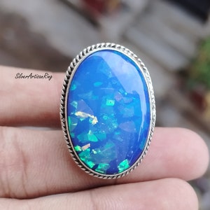 Könnte beinhalten: Ein silberner Ring mit einem großen ovalen blauen Opal-Edelstein. Der Opal hat einen schimmernden, irisierenden Effekt mit grünen und goldenen Blitzen.