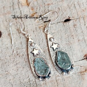 May include: A pair of silver teardrop-shaped earrings with a textured finish. Each earring features a light blue gemstone and a gold star.