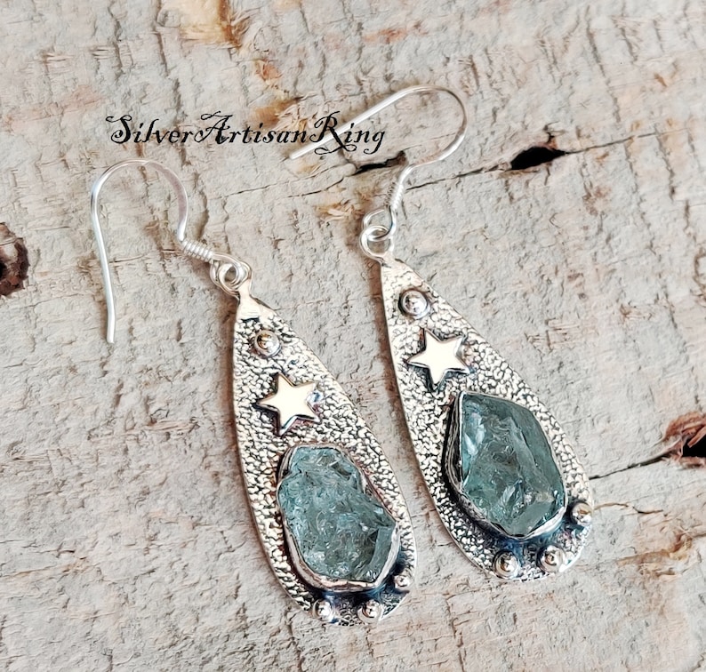 May include: A pair of silver teardrop-shaped earrings with a textured finish. Each earring features a light blue gemstone and a gold star.