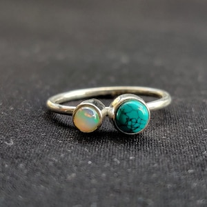 Könnte beinhalten: Ein silberner Ring mit zwei runden Steinen. Der Stein links ist ein weißer Opal, und der Stein rechts ist ein Türkis.