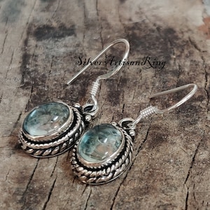 May include: Silver earrings with a blue gemstone set in a silver bezel with a rope-like design. The earrings have a hook closure.