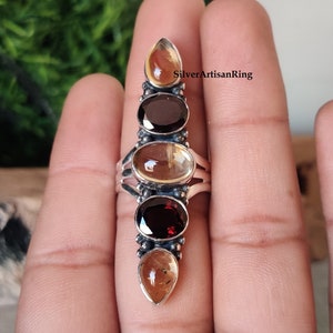 May include: A silver ring with five gemstones. The ring features a vertical row of alternating clear and red gemstones. The ring is on a finger.