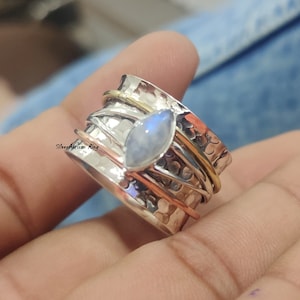 Puede incluir: Un anillo giratorio de plata con una piedra de luna y detalles dorados. El anillo está martillado y tiene un acabado texturizado.