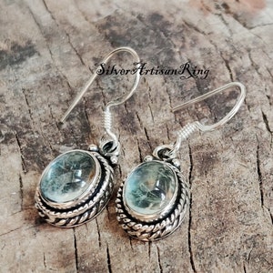 May include: Silver earrings with a light blue oval gemstone set in a silver bezel with a rope-like design. The earrings have a hook closure.