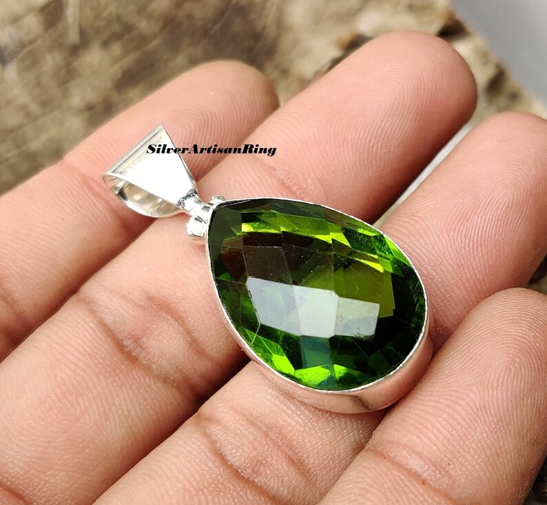 May include: A pear-shaped green gemstone pendant with a silver setting. The pendant is held in a hand.